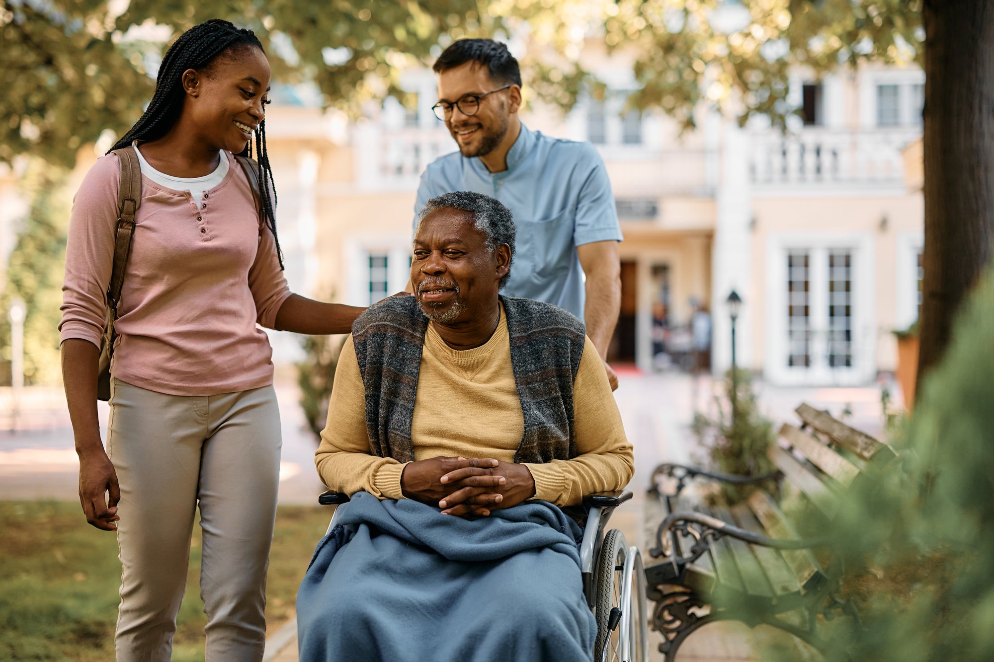 Two caregivers, one male and one female, smile while interacting with an elderly man in a wheelchair outside a residential building