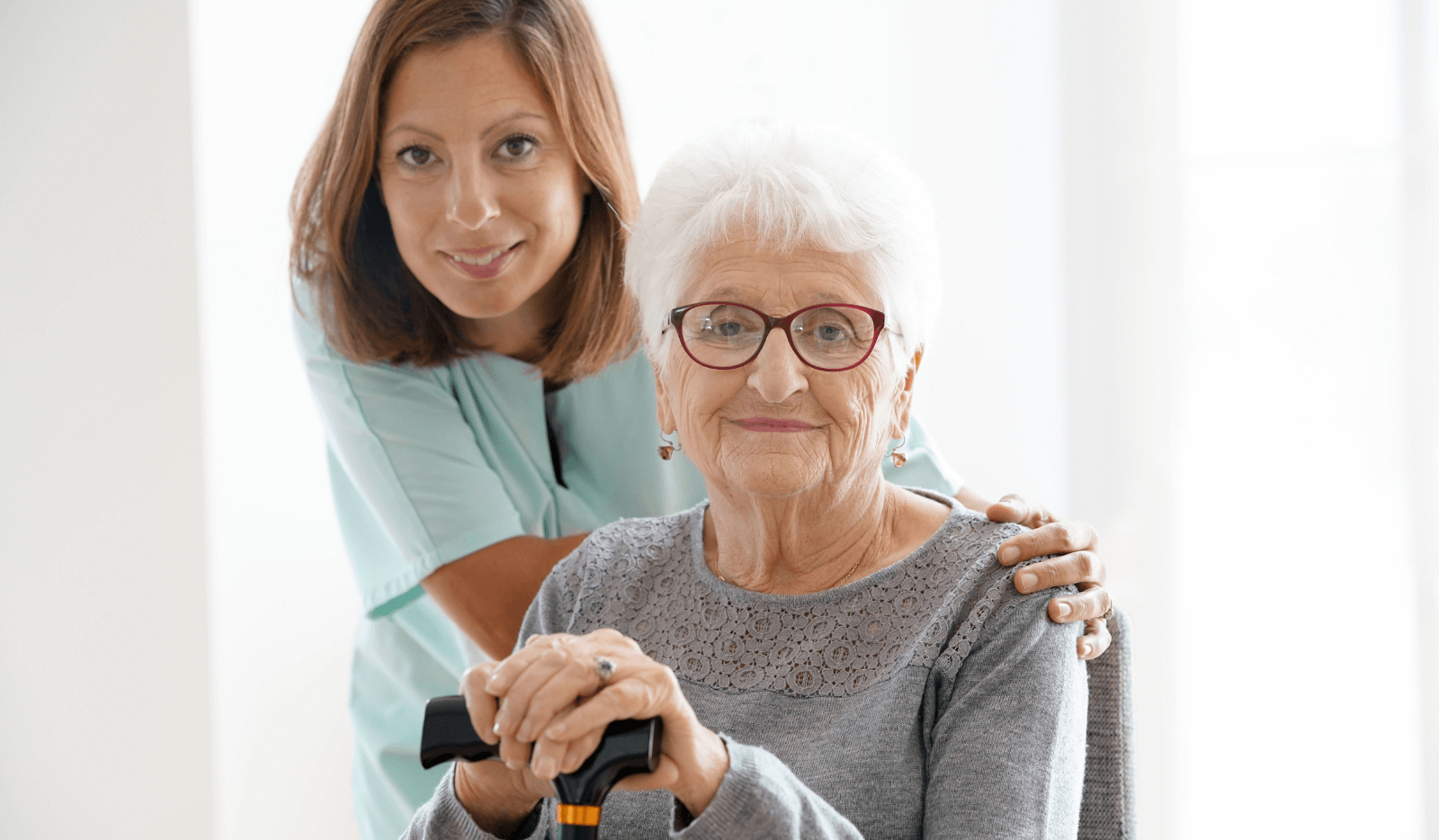 An elderly woman with glasses holds a cane while a younger woman in a teal uniform stands beside her, placing a hand on her shoulder.