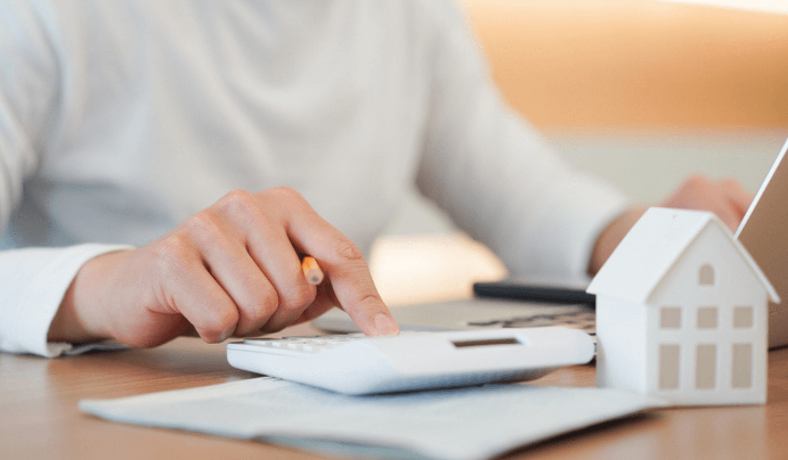 Hand pressing a calculator next to a small model house on a desk.