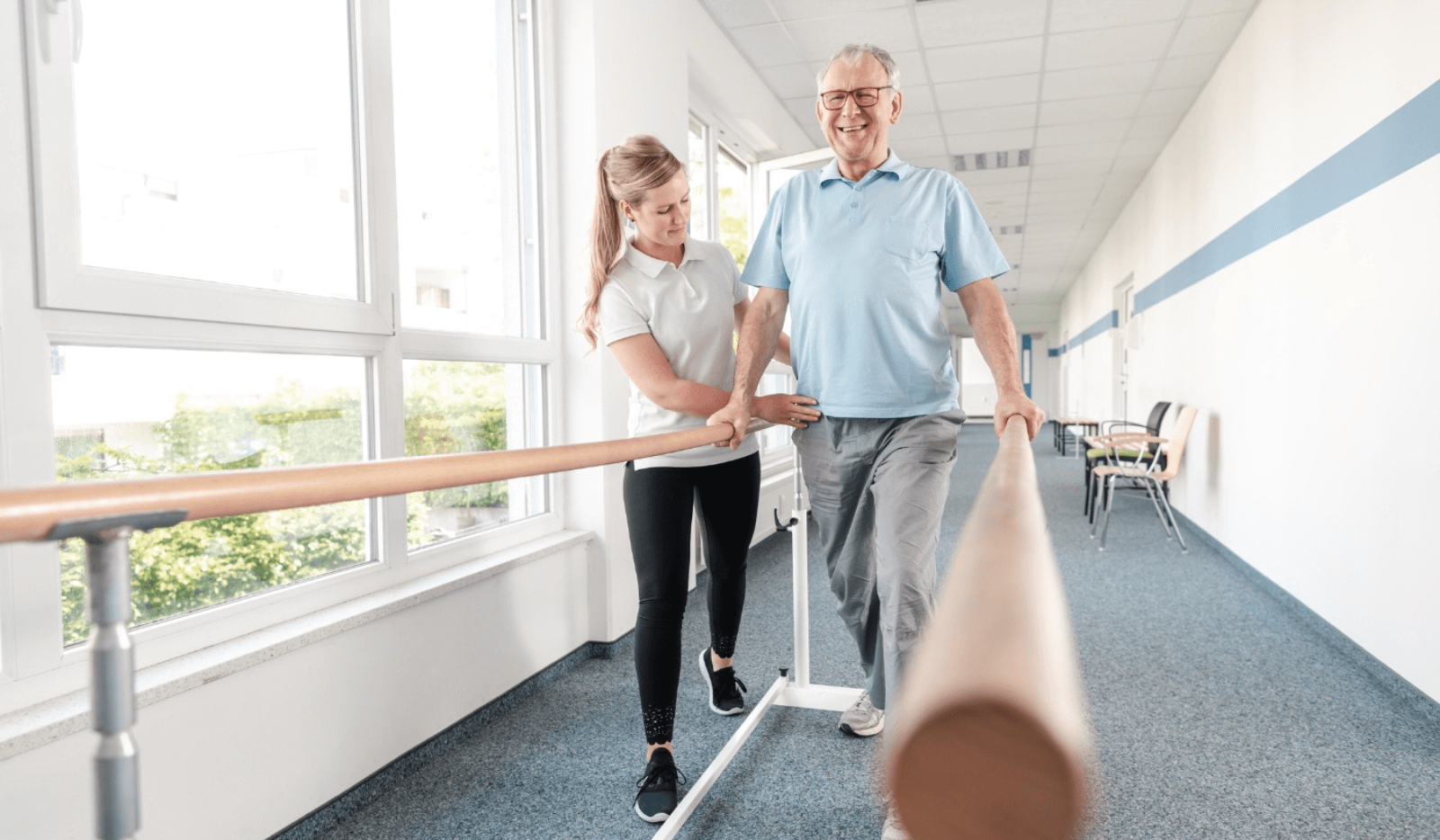 Elderly man walking with support on a handrail, assisted by a physical therapist in a bright corridor.