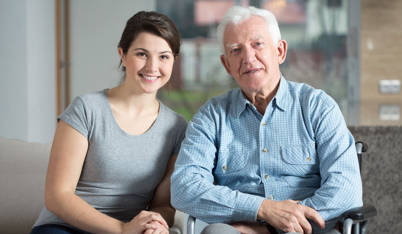 Young woman and older man smiling indoors, with the man seated in a wheelchair.